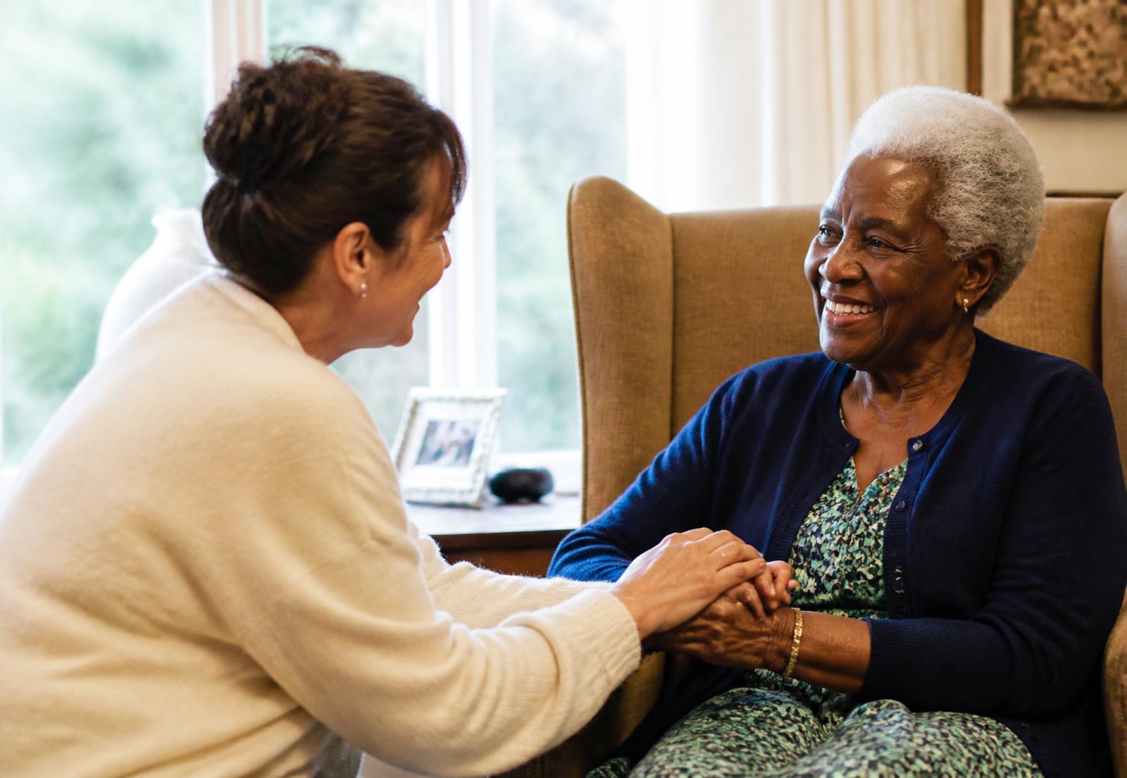 A caregiver holding the hand of a smiling elderly woman in her living room